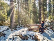 Frau sitzt auf Bank und blickt auf Sonnenstrahlen, die durch Bäume fließen — Stockfoto
