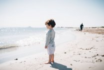 Vista lateral do bebê bonito de pé com os pés descalços na areia e olhando para o mar calmo brilhante na bela praia no dia ensolarado — Fotografia de Stock