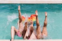 From above legs of barefoot women in swimwear holding LGBT flag and falling into swimming pool on resort — Stock Photo