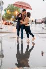 Весёлый молодой человек и женщина с зонтиком обнимаются и смотрят друг на друга, стоя на мокрой городской улице в дождливый день — стоковое фото