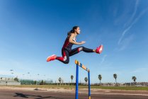Fuerte mujer joven en ropa deportiva saltando sobre el obstáculo contra el cielo azul durante el entrenamiento en el estadio - foto de stock