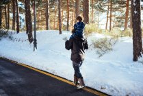 Вид сбоку на молодого человека в темной одежде, держащего ребенка в общей сложности на шее и идущего по желтой полосе по дороге вдоль снежного леса — стоковое фото