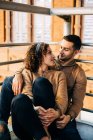 Fröhliche junge Mann und Frau umarmen und schauen einander an, während sie im beleuchteten Pavillon beim Date sitzen — Stockfoto