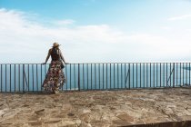 Woman in summer dress and hat standing at fence on stone terrace with seascape on background — Stock Photo