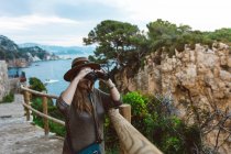 Woman observing view with binocular while standing at fence on stone walkway on seashore — Stock Photo