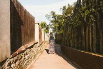Rückansicht einer nicht wiederzuerkennenden Frau mit Strohhut, die bei Sonnenschein auf einer schmalen Straße spaziert — Stockfoto