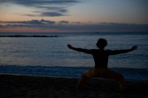 Rückenansicht eines sportlichen Mannes in gelben Hosen, der barfuß Yoga-Meditationstraining am Sandstrand am Abend des Sonnenuntergangs durchführt — Stockfoto