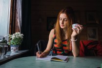 Mulher adulta pensando enquanto se senta à mesa no quarto aconchegante e escrevendo no caderno — Fotografia de Stock