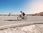 Healthy man riding bicycle on road in sunny day with barren landscape on background — Stock Photo