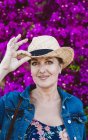 Woman wearing hat looking at camera while standing near pink flowers shrubs — Stock Photo