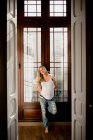 Mujer embarazada feliz en ropa casual de pie cerca de la ventana en casa - foto de stock