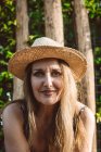 Довольна взрослая женщина в летней соломенной шляпе сидя против зеленых деревьев — стоковое фото