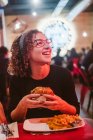 Mulher nova com fome comer hambúrguer saboroso enquanto sentado à mesa em café brilhantemente iluminado — Fotografia de Stock