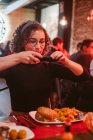 Jeune femme avec les cheveux bouclés en utilisant smartphone pour prendre des photos de hamburger et frites tout en étant assis à la table de café — Photo de stock