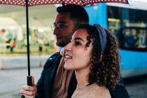 Вид збоку веселий молодий чоловік і жінка з парасолькою, що йде на мокрій міській вулиці в дощовий день — стокове фото