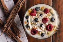 Сверху аппетитный ароматный йогурт с малиновым овсянкой из стекла и красными чашами, украшенными коричными палочками на деревянном фоне — стоковое фото