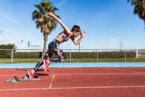 Сильная спортсменка в спортивной одежде быстро бегает против голубого неба в солнечный день на стадионе — стоковое фото