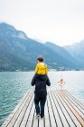 Back view of young man in dark clothes holding child in yellow jacket on neck and walking along wooden pier to calm water and breathtaking landscape in cloudy day — Stock Photo