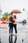Vista lateral de alegre joven hombre y mujer con paraguas abrazándose y mirándose mientras están de pie en la calle - foto de stock