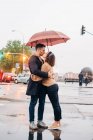 Seitenansicht von fröhlichen jungen Mann und Frau mit Regenschirm umarmen und einander anschauen, während sie auf der Straße stehen — Stockfoto