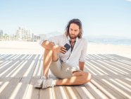 Barbudo teléfono inteligente de navegación masculina mientras está sentado en gazebo en la playa de arena cerca del mar en el día soleado - foto de stock