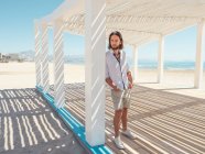 Красивый бородатый мужчина смотрит в сторону, опираясь на колонну белой беседки на песчаном пляже — стоковое фото