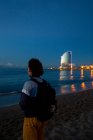 Back view of young man with backpack standing on sandy beach and looking along see and evening city — Stock Photo