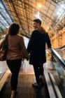 Вид сзади на молодого человека и женщину, держащихся за руки, стоя на движущейся дорожке во время свидания в ярко освещенном торговом центре — стоковое фото