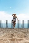 Жінка в літній сукні і солом'яному капелюсі, що стоїть на паркані на кам'яній терасі з морським пейзажем на фоні — стокове фото