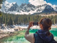 Vista posteriore della giovane donna in giù gilet tiro al telefono maestoso paesaggio di acqua turchese circondato da montagne rocciose e foresta in giornata nuvolosa — Foto stock
