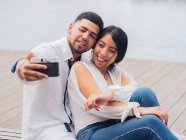 Jovem casal sentado e se divertindo tirando uma foto selfie no banco de madeira no litoral arenoso e beijando — Fotografia de Stock