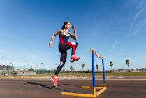 Strong young woman in sportswear leaping over hurdle against blue sky during workout on stadium — Stock Photo
