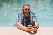 Взрослый мужчина с бородой в стильных оттенках и рубашке, просматривающий смартфон, расслабляясь в чистой воде бассейна — стоковое фото