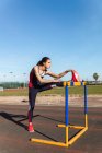 Strong young woman in sportswear stretching over hurdle against blue sky during workout on stadium — Stock Photo