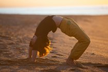 Hombre deportivo en pantalones amarillos descalzo realizando ejercicios de meditación de yoga en la playa de arena al atardecer - foto de stock