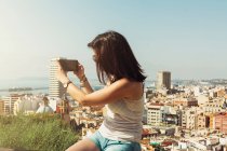 Vista lateral de una joven en camiseta blanca tomando fotos de las vistas de la ciudad desde el balcón en un día luminoso en Alicante España - foto de stock