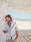 Bonito homem barbudo usando smartphone enquanto se inclina no pilar de gazebo branco na praia arenosa — Fotografia de Stock