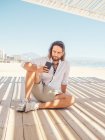 Smartphone de navegação masculino barbudo enquanto sentado no gazebo na praia de areia perto do mar no dia ensolarado — Fotografia de Stock