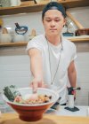 Joven cocinero masculino poniendo en el mostrador gran tazón con delicioso plato japonés llamado ramen en la cafetería oriental - foto de stock