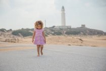 Кудрявый ребенок в полосатой ходьбе по дороге песчаного пляжа на размытом природном фоне — стоковое фото