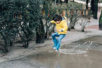 Захоплений хлопчик в пальто і гаманець розважається на вулиці і стрибає на калюжі — стокове фото
