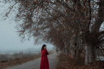 Femme rêveuse en robe rouge marchant le long de la route vide de terrain mystérieux brumeux — Photo de stock