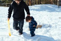 Молодий чоловік в темному теплому одязі тримає руки з дитиною в смугастому комбінезоні і ходить снігом в сонячний день — стокове фото