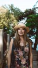 Доросла жінка в солом'яному капелюсі і квітковій сукні дивиться на камеру на сонячній вулиці — стокове фото
