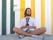 Bärtiger Mann in lässigem Outfit hält Smartphone in der Hand, während er auf Sand an gestreifter Wand im Resort sitzt — Stockfoto