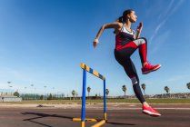 Starke junge Frau in Sportbekleidung springt beim Training im Stadion über Hürde gegen blauen Himmel — Stockfoto
