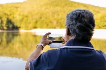 Vista trasera de un hombre mayor tomando fotos con teléfono móvil mientras contempla el increíble paisaje de la montaña del lago - foto de stock