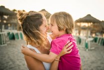 Visão lateral da mulher rindo carregando alegre filho brincalhão a mãos enquanto esfregando narizes na praia ao pôr do sol — Fotografia de Stock