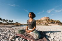 Attractive young woman meditating and praying wearing japamala collar in lotus yoga posture on sandy beach in bright day — Stock Photo