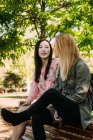 Две многорасовые молодые женщины в повседневных нарядах разговаривают и смотрят друг на друга, сидя на скамейке в парке — стоковое фото